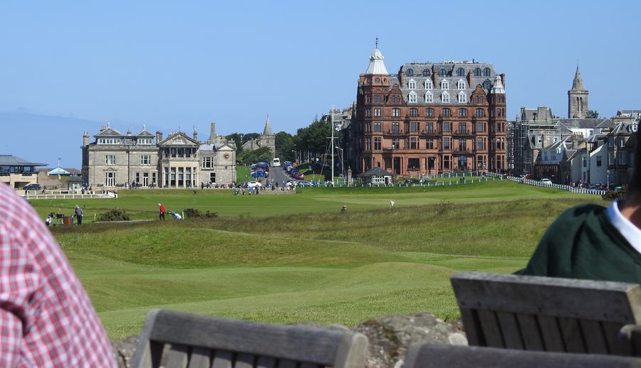 A green golf links in the foreground, a low pale clubhouse to the left and a tall red sandstone building with turrets to the right, spectators standing along the edge.