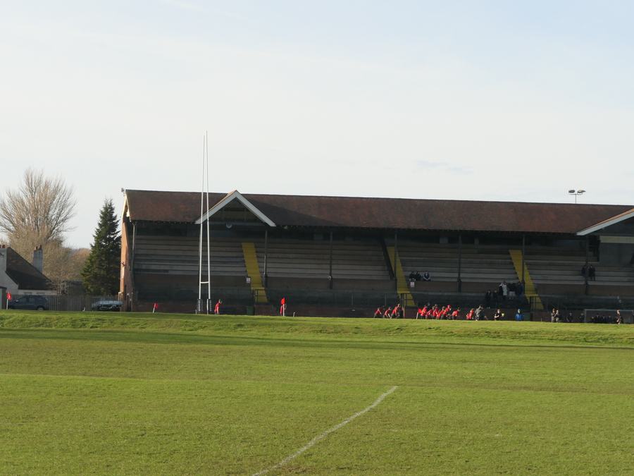 A long low grandstand with a pitched roof on stanchions, facing empty green rugby pitches with white markings.