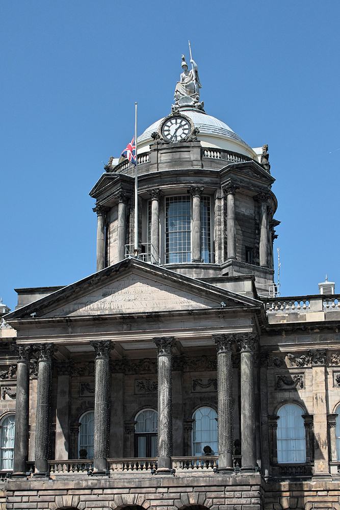 A Georgian civic building of pale stone with a Corinthian portico below a drum and dome carrying a clock and a statue, against a blue sky.