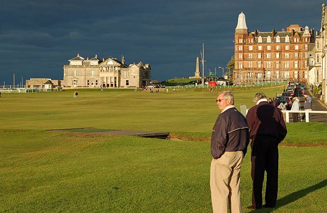 A sunlit golf fairway running towards a pale stone clubhouse and a large red sandstone building, both bright against a near-black storm sky.