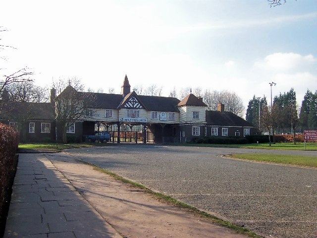 A long black-and-white timbered pavilion building set behind a broad empty tarmac forecourt, with a paved footpath running along the left.