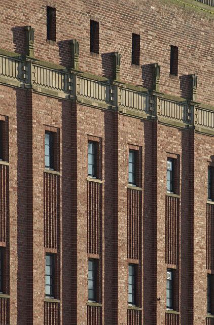 Close view of a brick facade with vertical stone-framed window strips and a banded parapet, in raking light.