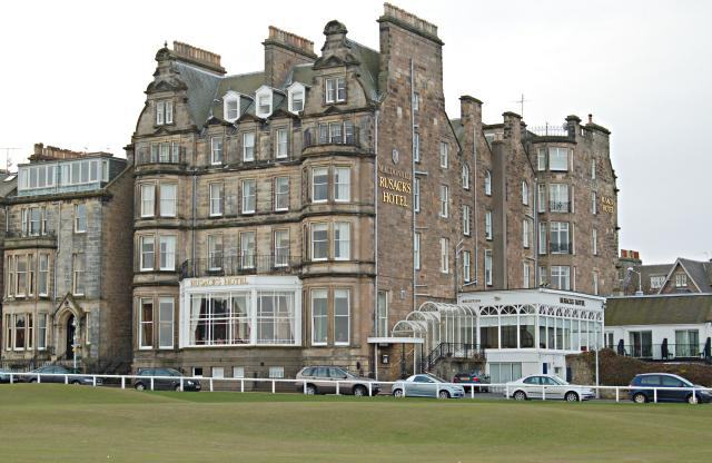 A five-storey grey stone hotel with dormers and a glazed conservatory along the front, seen across a strip of golf links with parked cars in between.
