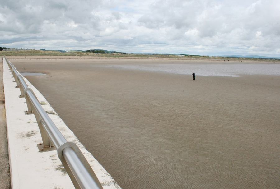 A very wide flat empty beach under a grey cloudy sky, a metal handrail along a concrete wall in the foreground and a single distant figure.