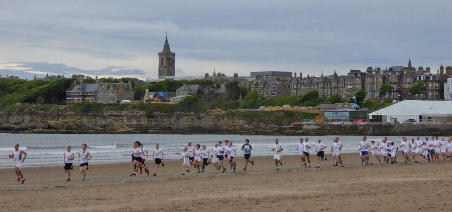 About twenty runners in white shirts and shorts spread across wet sand at the water's edge, with the town and its church tower on the low cliff behind.