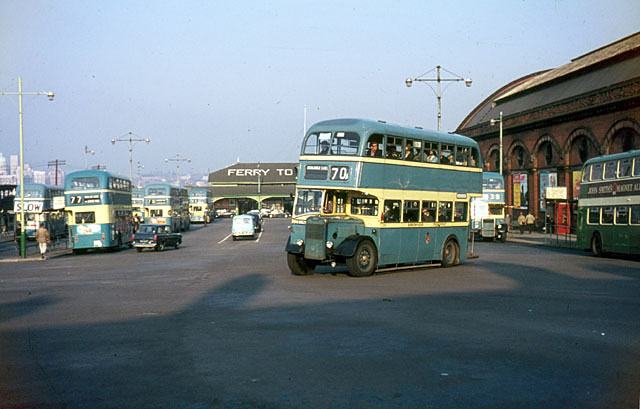 A colour photograph of 1960s double-decker buses in blue and cream standing on a wide tarmac apron in front of a brick terminal building with FERRY TO signs.
