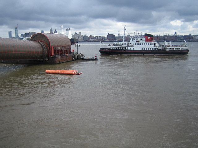 A ribbed metal covered walkway sloping down to a floating landing stage on a grey river, a white and black ferry alongside and a city waterfront on the far bank.