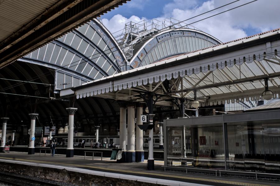 A great curving glazed iron roof over railway platforms, carried on slender columns with decorative spandrels, a platform 6 sign in the middle distance.