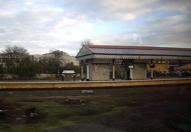 The far end of a railway station seen from a train, with a canopy on cast iron columns, low buildings and bare trees under a grey sky.