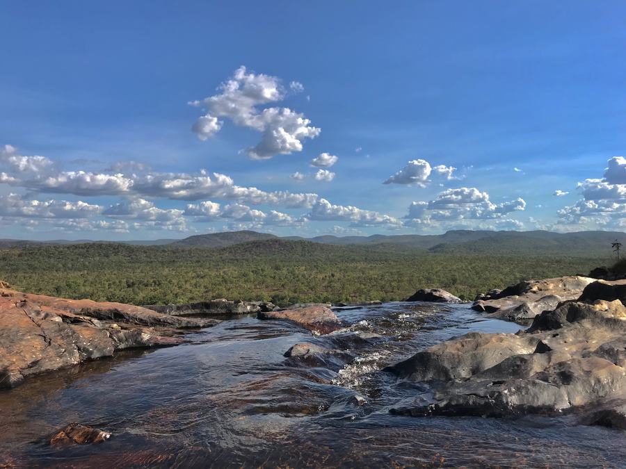 Shallow rock pools on a flat sandstone shelf running to the edge of a cliff, an enormous view of green bushland and low ranges beyond.