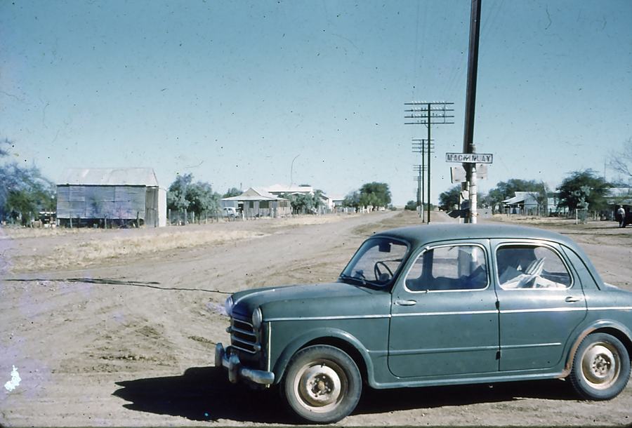 A pale blue 1950s sedan parked on a wide dirt road, low iron-roofed buildings and power poles either side.