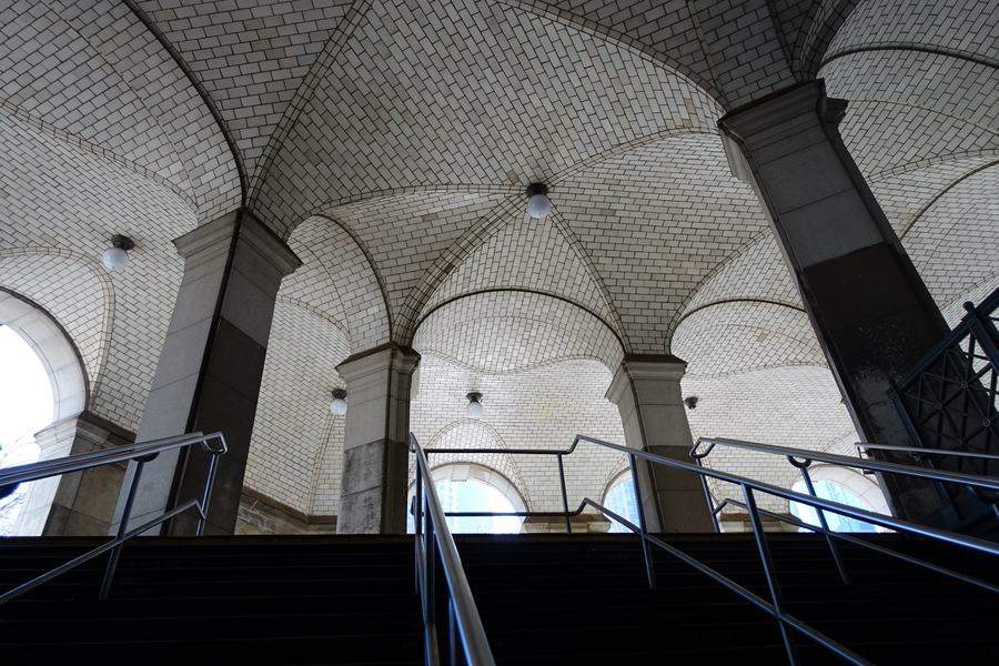 Looking up a subway stairway into a high vaulted hall lined with small white tiles, heavy stone piers, daylight through arched openings.