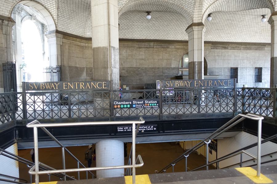 A white tiled vaulted arcade with the words SVBWAY ENTRANCE carved above two stairways leading down, iron railings, a Chambers Street Station sign.