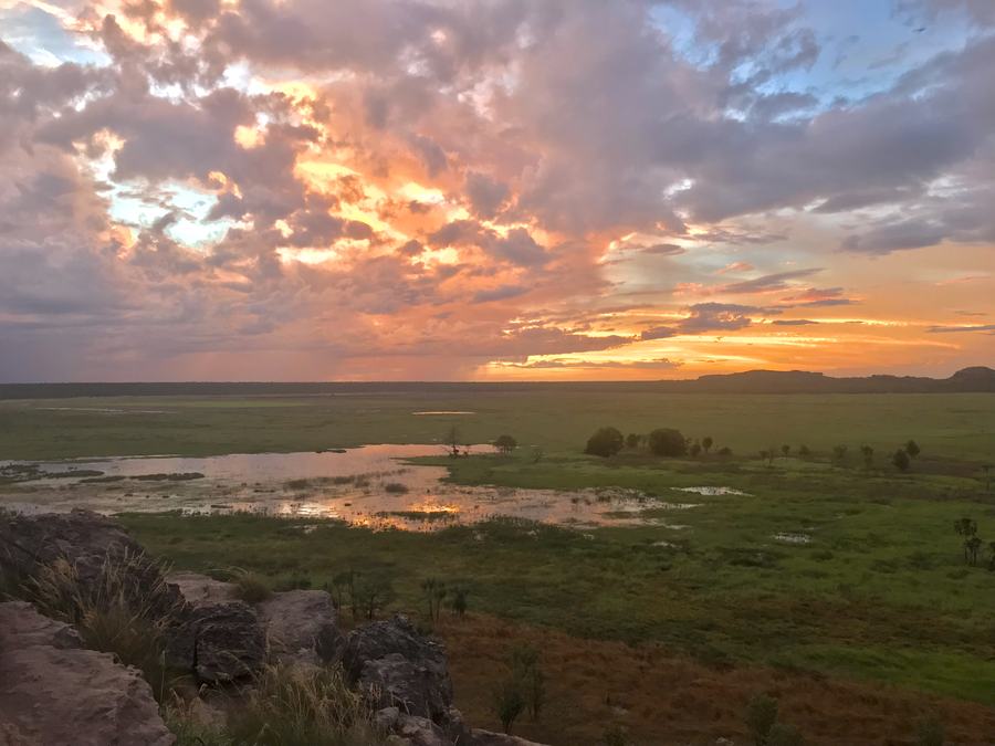 A wide flat green floodplain under a high orange and grey sunset sky, seen from a rock ledge in the foreground.