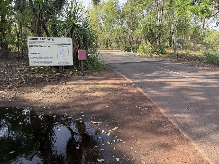 A weathered white metal sign beside a bush track headed UBIRR ART SITE, RESTRICTED ENTRY, with opening times and a small map panel.