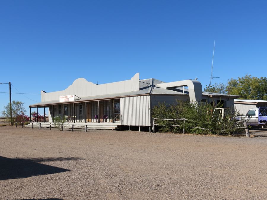 A single-storey white pub with a deep verandah on a bare red apron, water tank and utility vehicles at the side, hard blue sky.