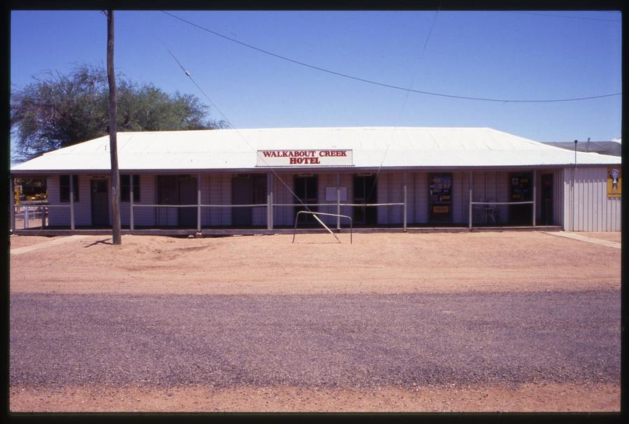 A long low pub with a corrugated roof and a WALKABOUT CREEK HOTEL sign, standing on bare reddish ground.