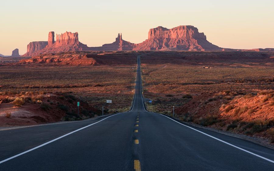 Empty two-lane highway running straight towards red sandstone buttes at sunrise