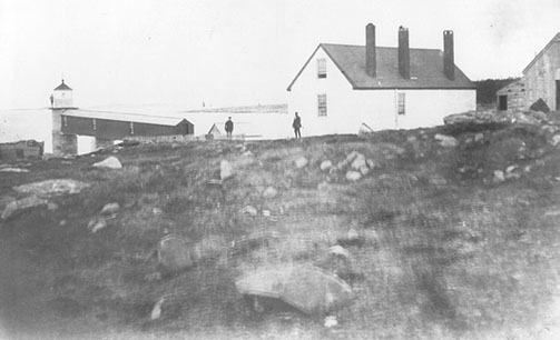 Old black and white photograph of the light station with two figures standing on the rocks