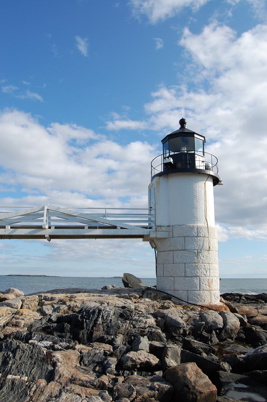 Short white lighthouse tower on a granite base with the walkway behind it