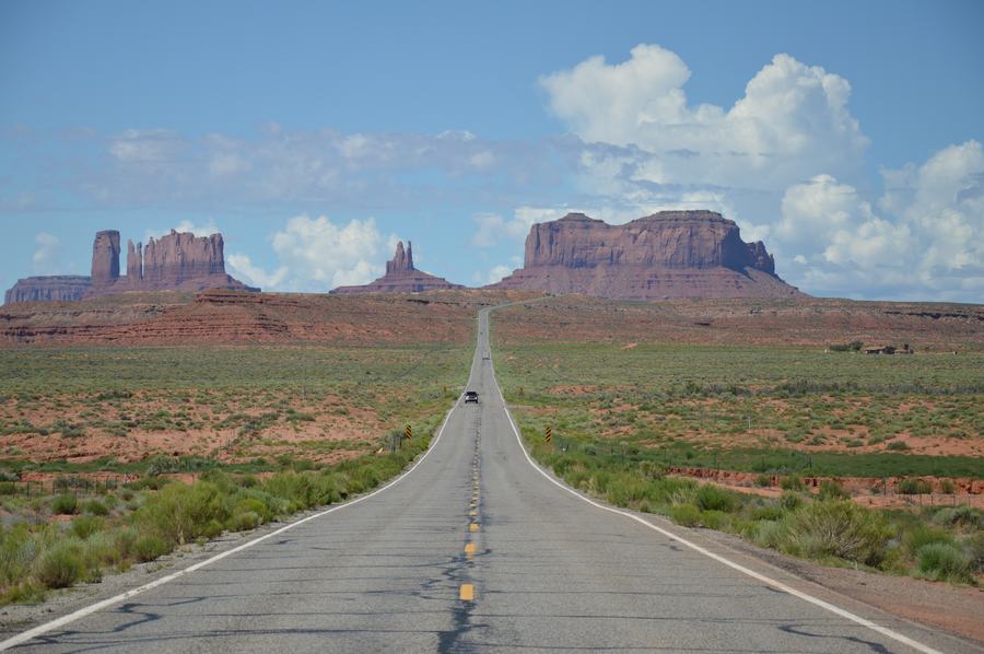Two-lane desert highway with a car in the distance and buttes on the skyline