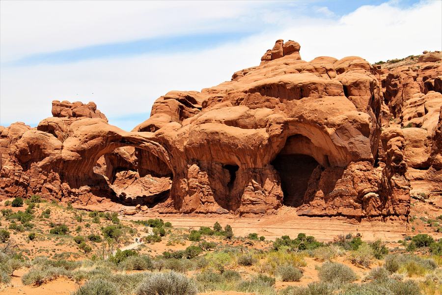A wide view of red rock cliffs with a natural arch at left and two deep alcoves at right, desert scrub in front and a footpath with small figures.