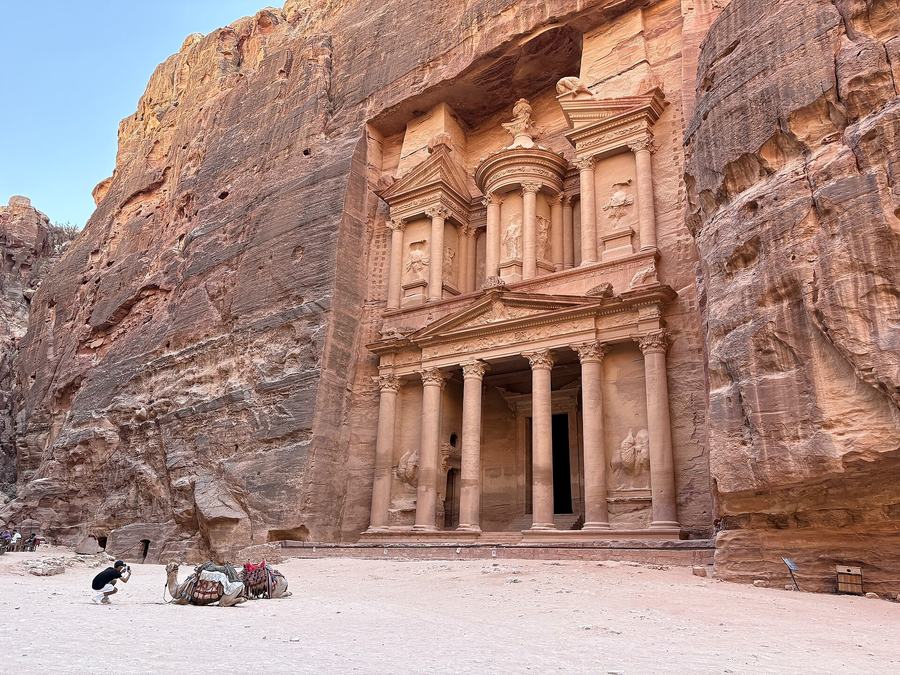 The full two-storey rock-cut facade of Petra's Treasury in daylight, with saddled camels resting on the sand in front and a person photographing them.