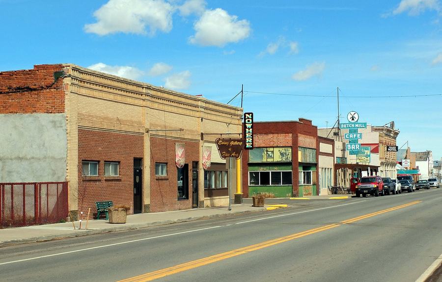 A small-town main street of low brick storefronts, several vacant, with hand-painted signs for an art gallery, a cafe and a hotel, and a two-lane road in front.