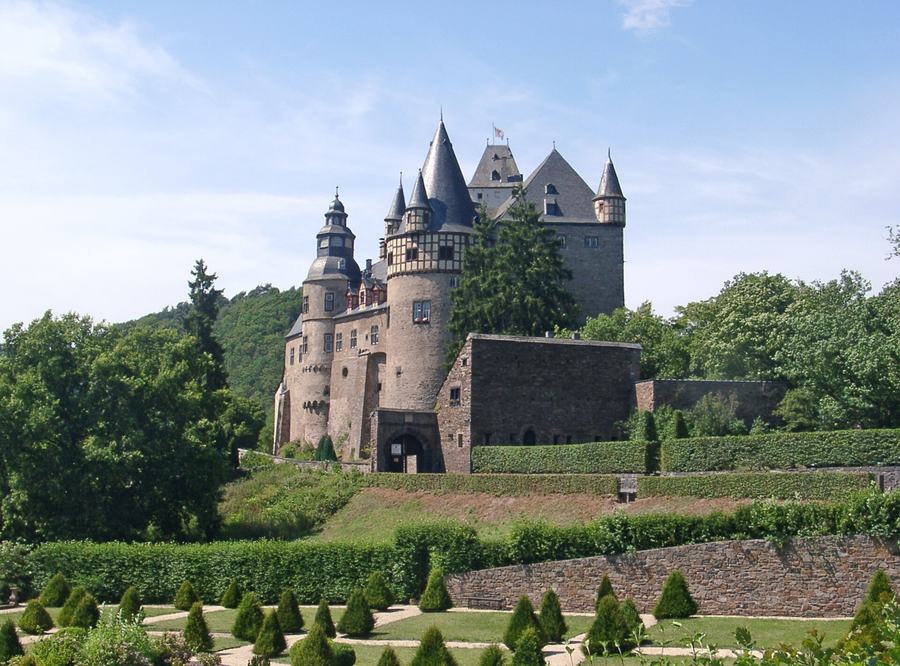 A hillside castle with conical slate roofs, round towers and half-timbered oriels, above a clipped hedge garden with cone-shaped shrubs.
