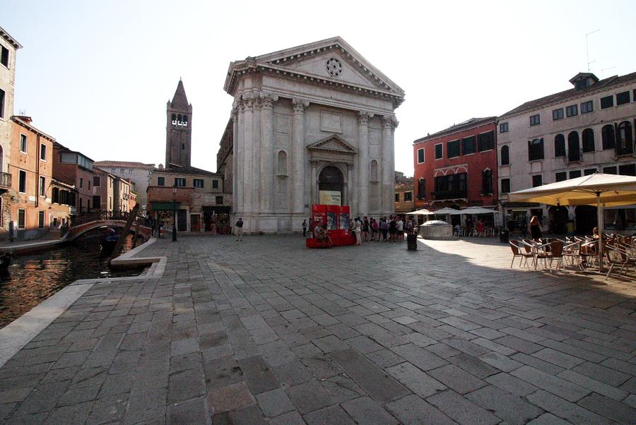 A wide paved Venetian square with a columned church front at the far end, café tables under umbrellas on one side and a canal with a small bridge on the other.