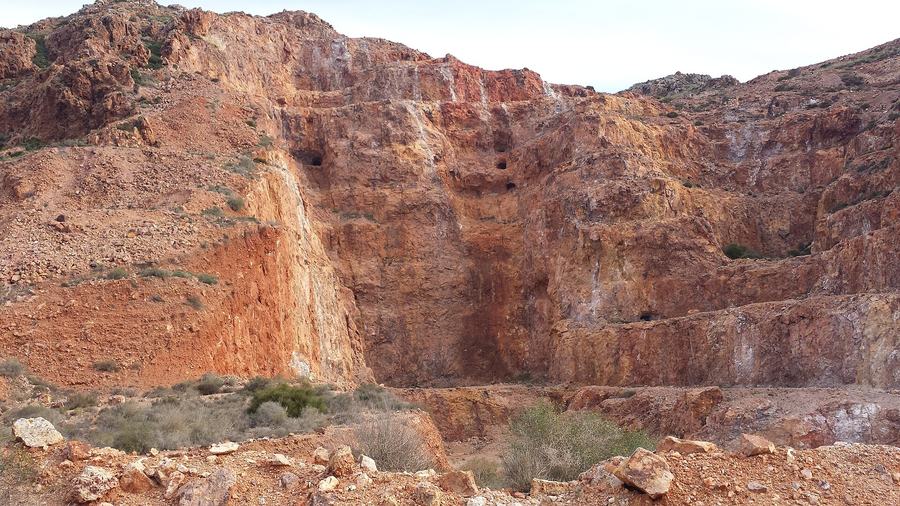 A stepped opencast face of orange and yellow rock with several small dark tunnel mouths, dry scrub in the foreground.