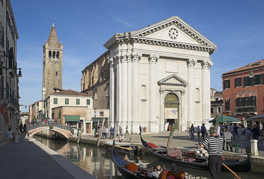 A white neoclassical church front with four columns and a pediment, seen across a canal in Venice; gondolas in the foreground and a tall brick bell tower behind.