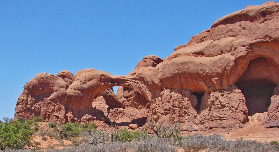 A red sandstone formation with a slender natural arch on the left and a large shadowed alcove eroded into the rock face on the right.