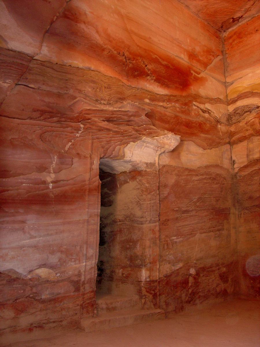 A plain rock-cut chamber with rippled red and cream sandstone walls and a rectangular doorway leading into a darker inner room.