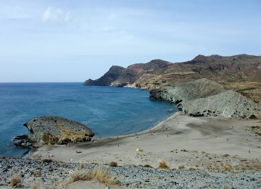 A curved sandy beach on a bare volcanic coast, with a large domed rock standing in shallow blue water at the near end.