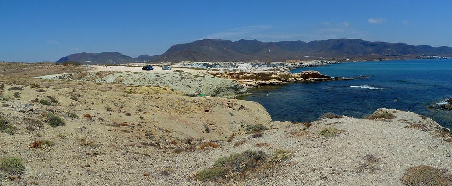 A panoramic view of a low rocky coastline of pale fossil dune stone beside deep blue sea, with brown mountains behind and two parked cars.