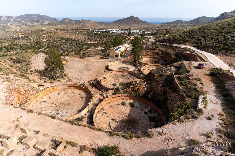 Looking down on a group of large empty circular concrete tanks in dry hills, with a road, a low modern building, a white village and the sea in the distance.
