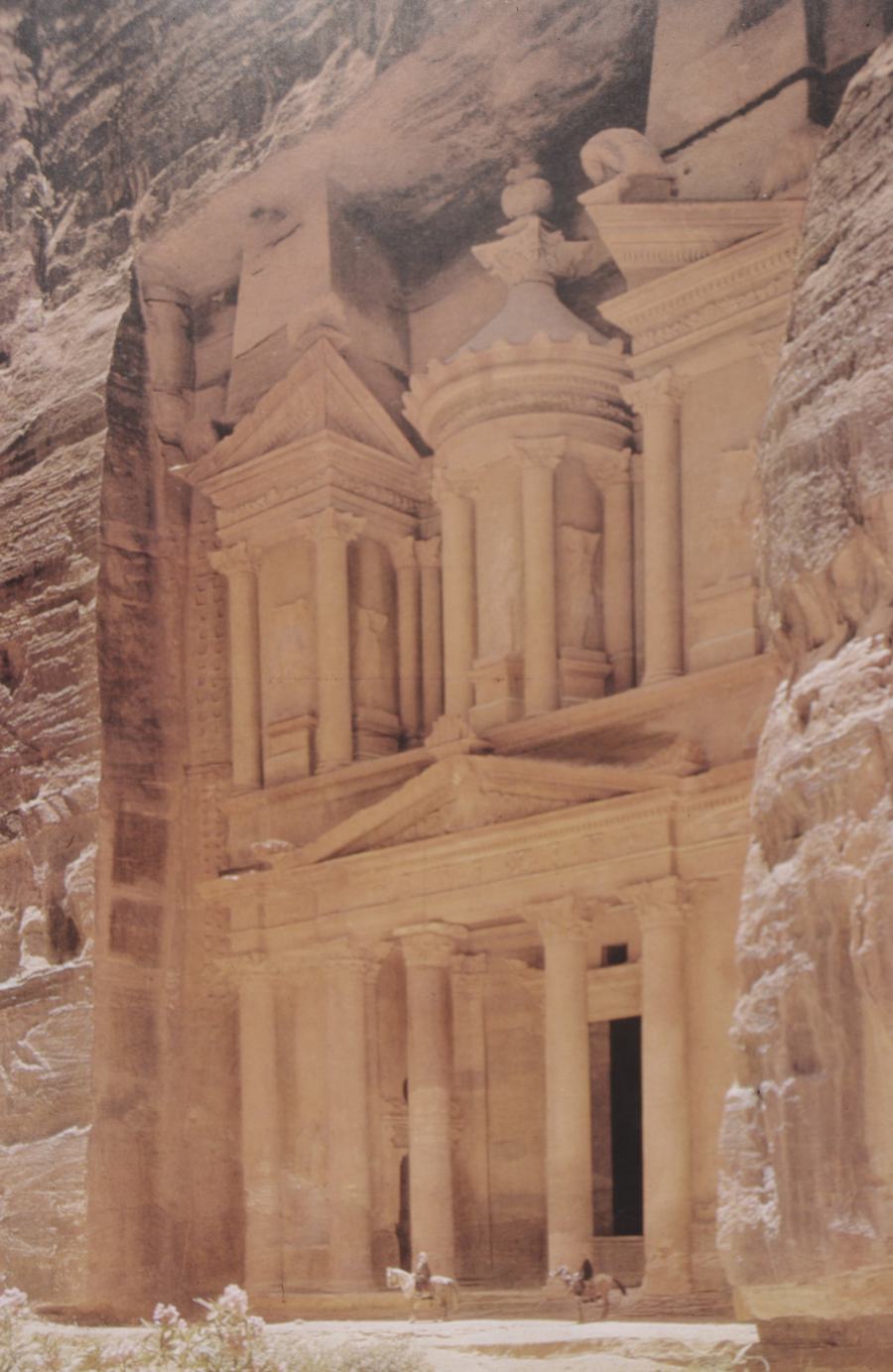A carved classical facade in pink sandstone framed by the dark walls of a narrow rock gorge, with two tiny figures at its base.