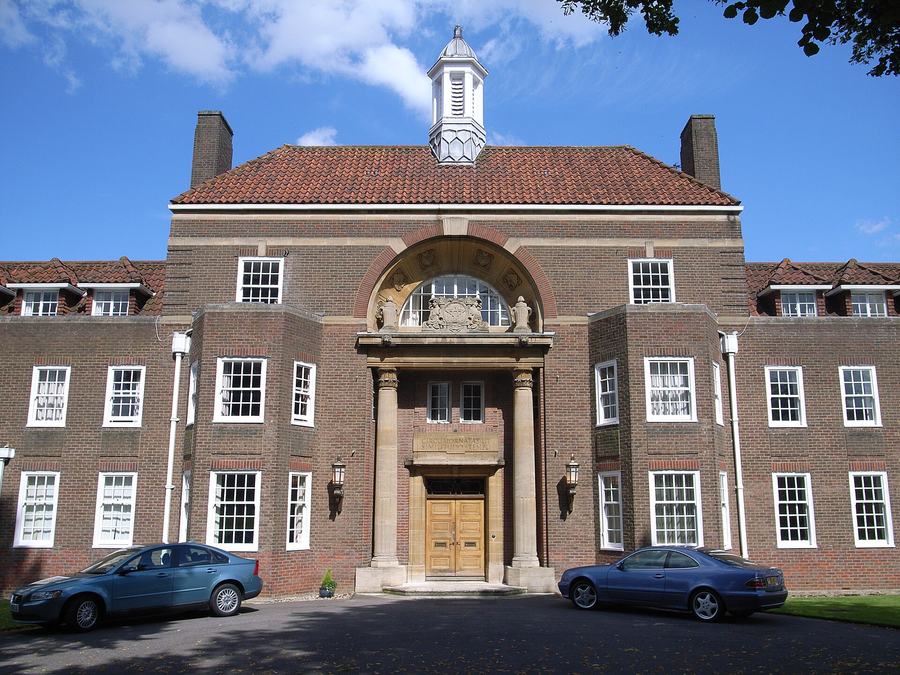 A symmetrical brown-brick school building of the 1930s with a white cupola, an arched window over a stone porch, and cars parked in front.