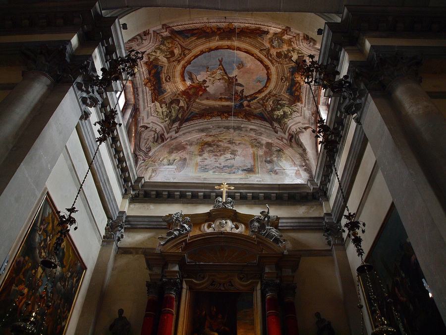 Looking up inside a church at an oval ceiling fresco of figures and clouds framed by gilded stucco, between two grey stone columns.