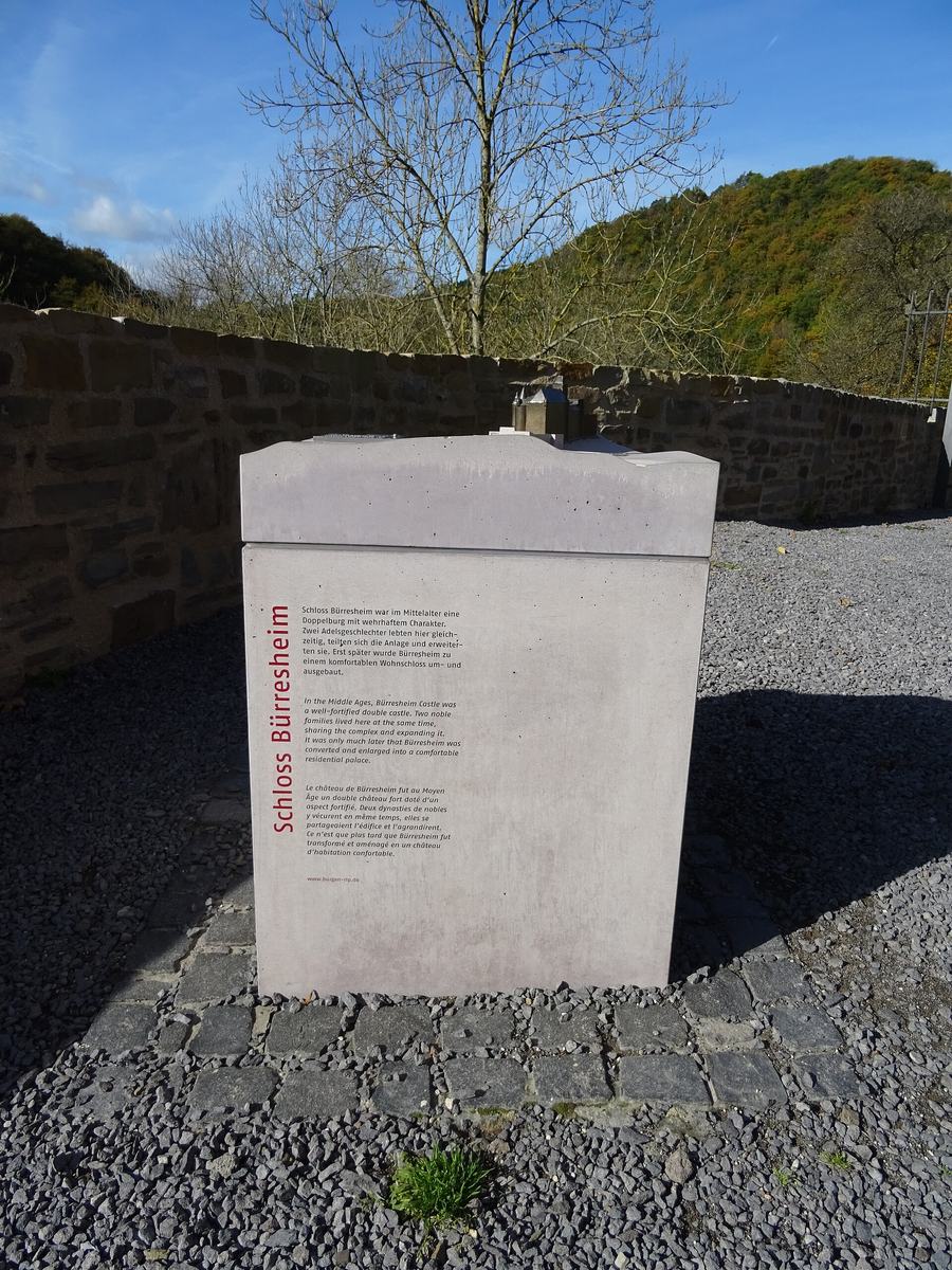 A pale concrete information block in a gravelled courtyard, carrying text in three languages and a small bronze relief model of the castle on its top.