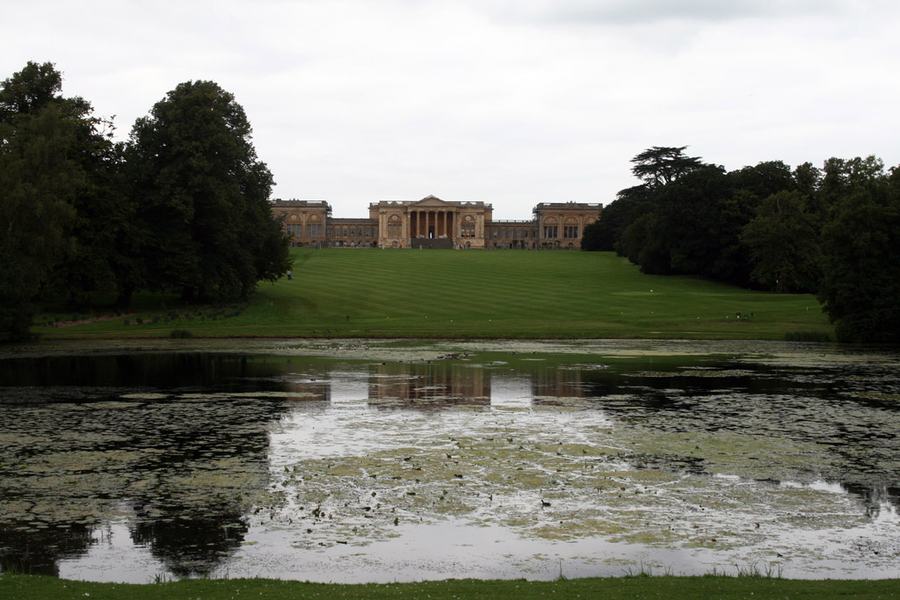 A long classical mansion with a columned central portico seen from across a weed-covered lake and a broad sloping lawn.