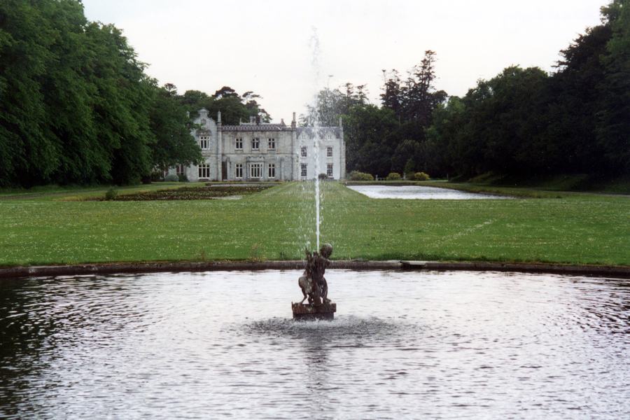 Killruddery House seen down the length of its formal water garden: a pale grey Elizabethan-revival house with gabled bays at the head of a long lawn, twin still canals behind, and in the foreground a round pond with a bronze figure and a jet of water rising high in front of the house.