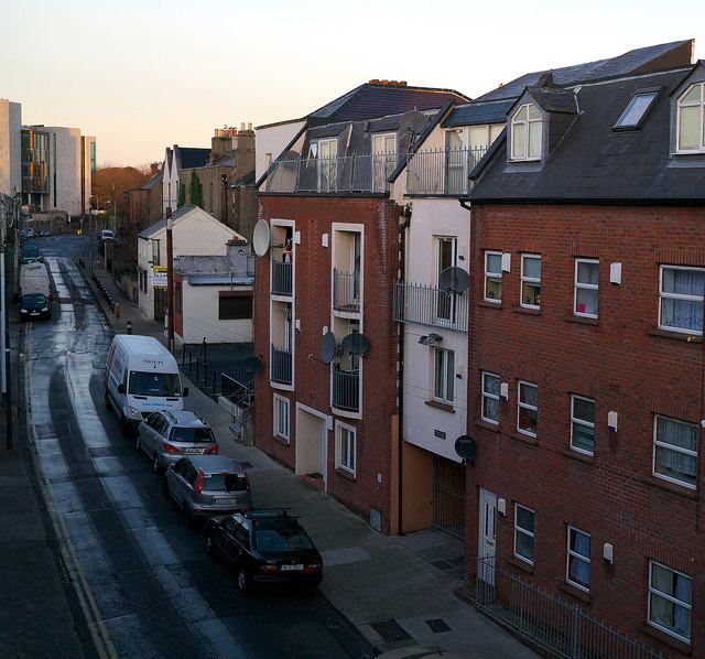 Montpelier Hill, Dublin, seen from above at dusk: a steep, wet street falling away to the left with cars and a white van parked along it, low white cottages on the far side, and a four-storey red-brick apartment block with satellite dishes and balconies filling the right of the frame.
