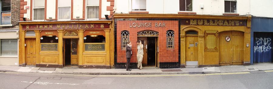 A wide, letterbox-shaped view of the whole frontage of Mulligan's pub on Poolbeg Street, Dublin: the timber JOHN MULLIGAN shopfront on the left, a dark red LOUNGE BAR entrance in the middle with two people standing in the doorway, and a second gold MULLIGANS sign on the right.