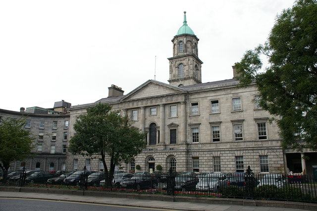 The granite front of the Rotunda Hospital on Parnell Street, Dublin: a long Georgian range with a pedimented centre and a green copper cupola on a square tower above it, railings and parked cars along the pavement in front.