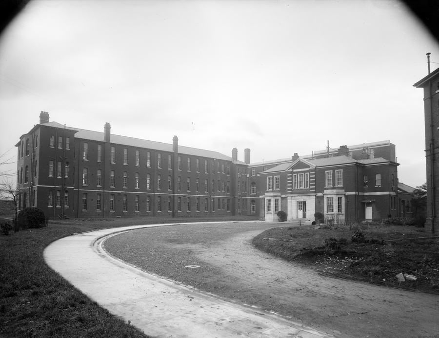 A black-and-white photograph of a large brick institutional building: a long three-storey ward block with rows of identical windows and tall chimneys stretching away to the left, a lower two-storey block with a pedimented centre on the right, and a curving gravel drive and clipped grass in front.