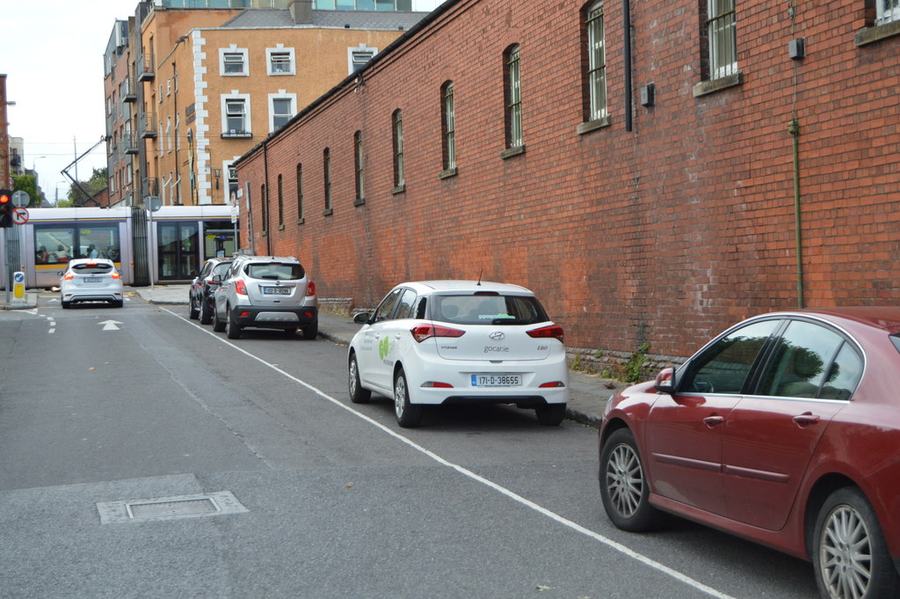 Temple Street West in Dublin 7, looking down the hill: parked cars along a kerb on the right and a long red-brick wall with tall arched windows running the length of the street, a Luas tram crossing at the bottom.