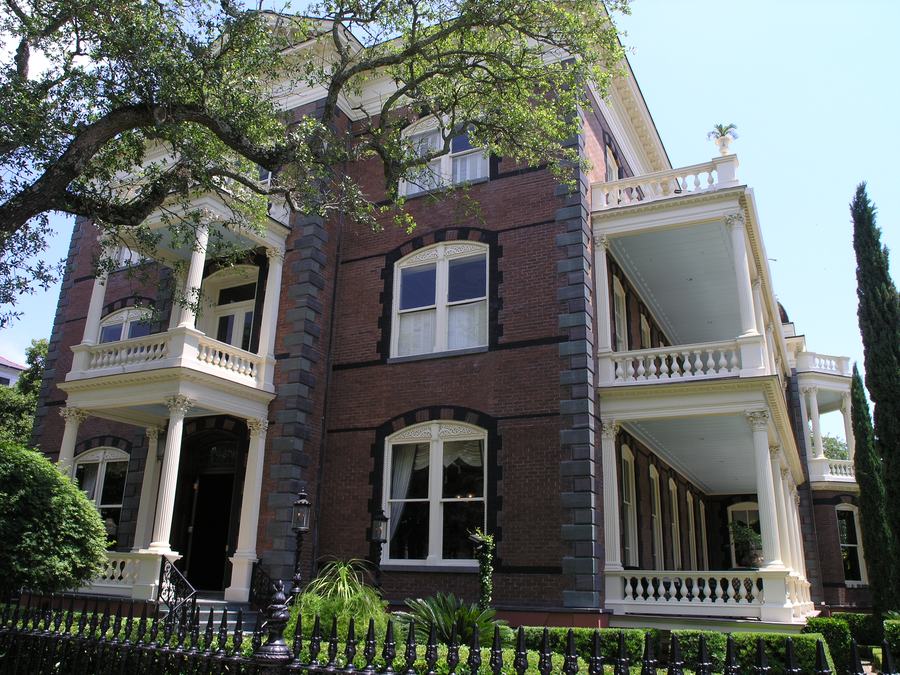 A tall ornate Victorian brick mansion behind a fence on a Charleston street, with arched windows and a pillared porch.