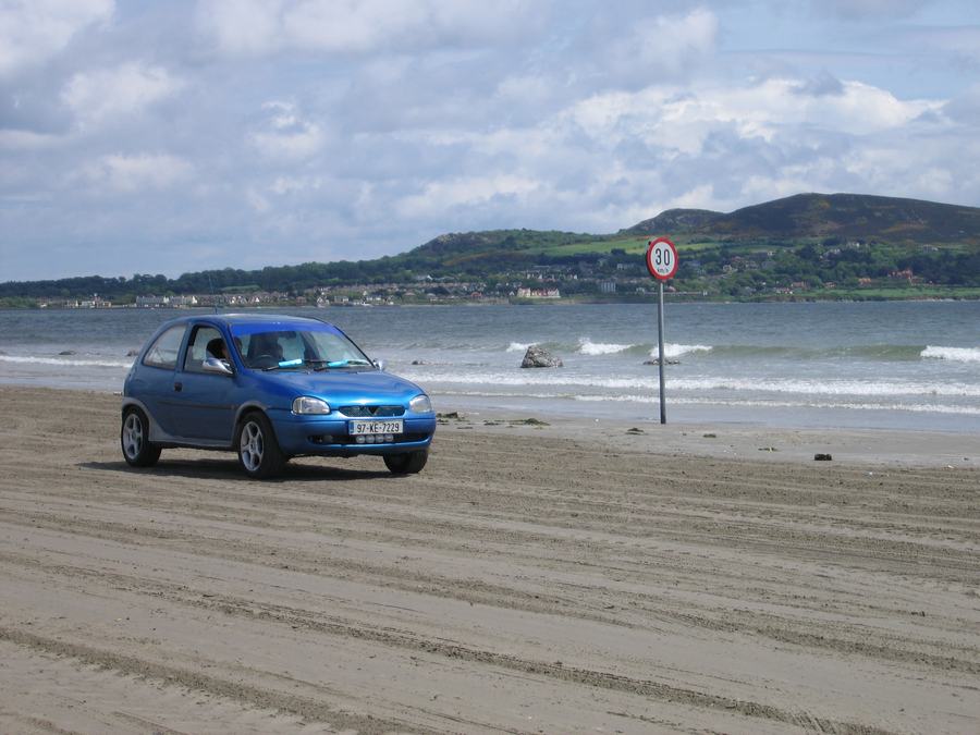 A small blue car parked on the hard sand of Dollymount Strand, Dublin, with the sea and the Howth headland behind it.